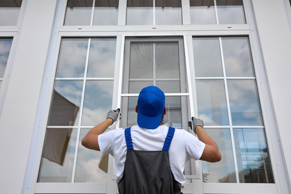A worker in a blue cap and gloves inspects a large window with multiple panes against a white frame. While assessing potential roof repair needs in New Castle County, DE, the clear glass reflects an overcast sky as the person is seen from behind, intently focused on their task.