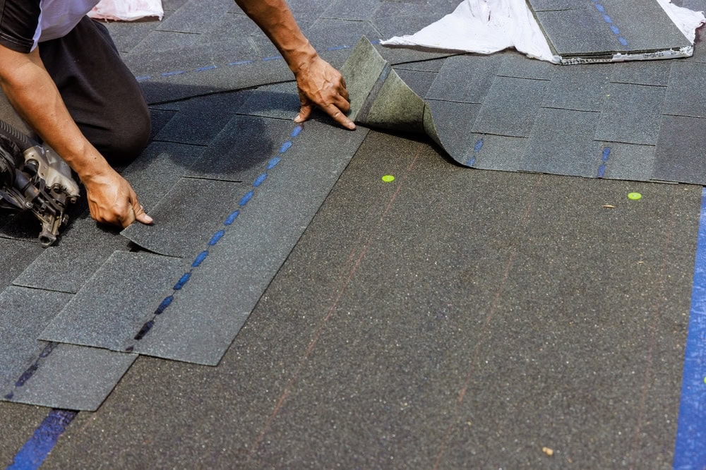 A skilled roofer in New Castle County is laying dark asphalt shingles on a roof. With precision, they use a nail gun to secure each shingle while their hands meticulously adjust the placement. The textured shingles align perfectly with the blue guidelines on the roof.