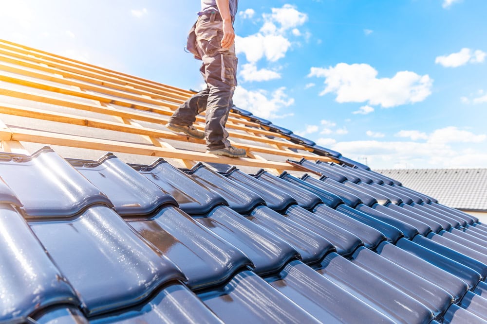 A person stands on a roof under construction in New Castle County, clad in workwear. One side of the roof boasts shiny blue tiles, while wooden beams await completion on the other. Overhead, a bright sky with scattered clouds stretches across the scene.