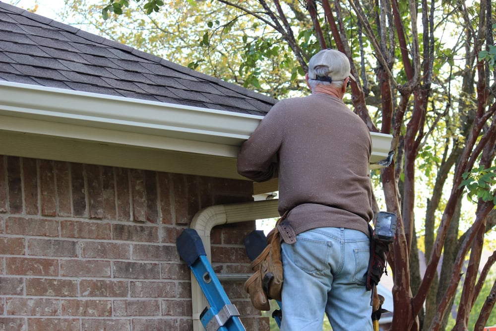 A roofer in a cap and brown sweater is perched on a ladder beside a brick building in New Castle County, DE, diligently working on a gutter. Equipped with a tool belt around their waist, they are framed by the silhouette of a tree in the background.