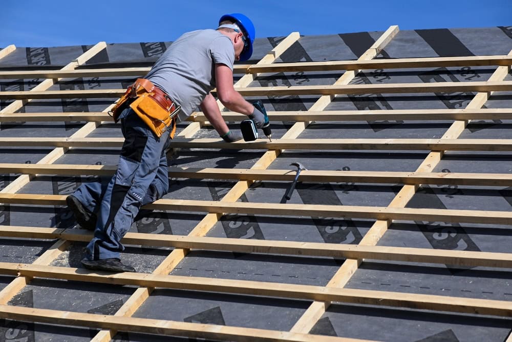 A skilled roofer in New Castle County, wearing a hard hat and tool belt, diligently works on a sloped roof under the clear blue sky. Dressed in a gray shirt and dark pants, they use a power drill to secure beams, showcasing their expertise in roof repair.