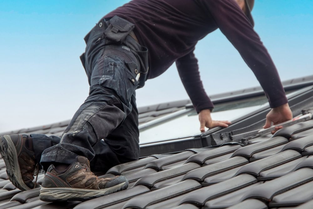 A roofer in dark clothing and hiking boots is kneeling on a textured, brown-tiled roof in New Castle County, DE, adjusting a skylight window. The sky in the background is clear and blue.