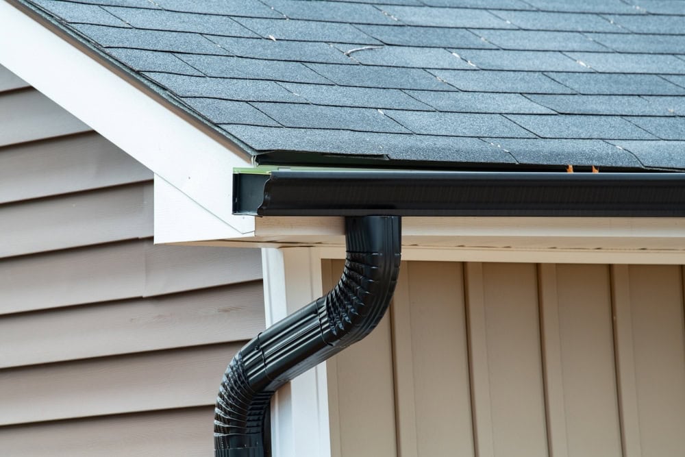 Close-up of a house roof corner in New Castle County, DE, featuring gray asphalt shingles, white eaves, and a black downspout. The beige siding highlights the architectural details crafted by a skilled roofer.