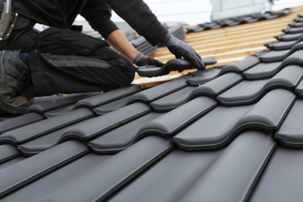 A skilled roofer in New Castle County dons gloves as they meticulously install black, wavy roof tiles on a sloped wooden structure. The image provides a close-up of the tiles and the artisan's hands, showcasing their expertise in roof repair throughout DE.