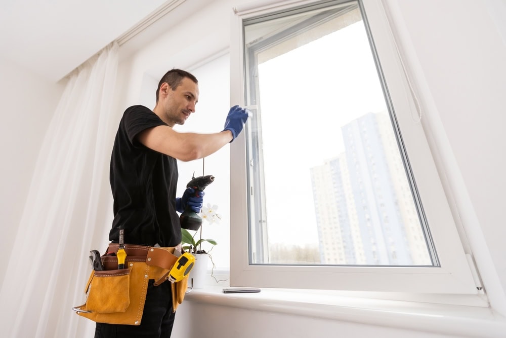 A man wearing a tool belt and gloves is expertly repairing a window indoors with a drill, much like he's done for countless roof repairs across New Castle County. A cityscape with tall buildings is visible outside, while a small white orchid graces the window sill.
