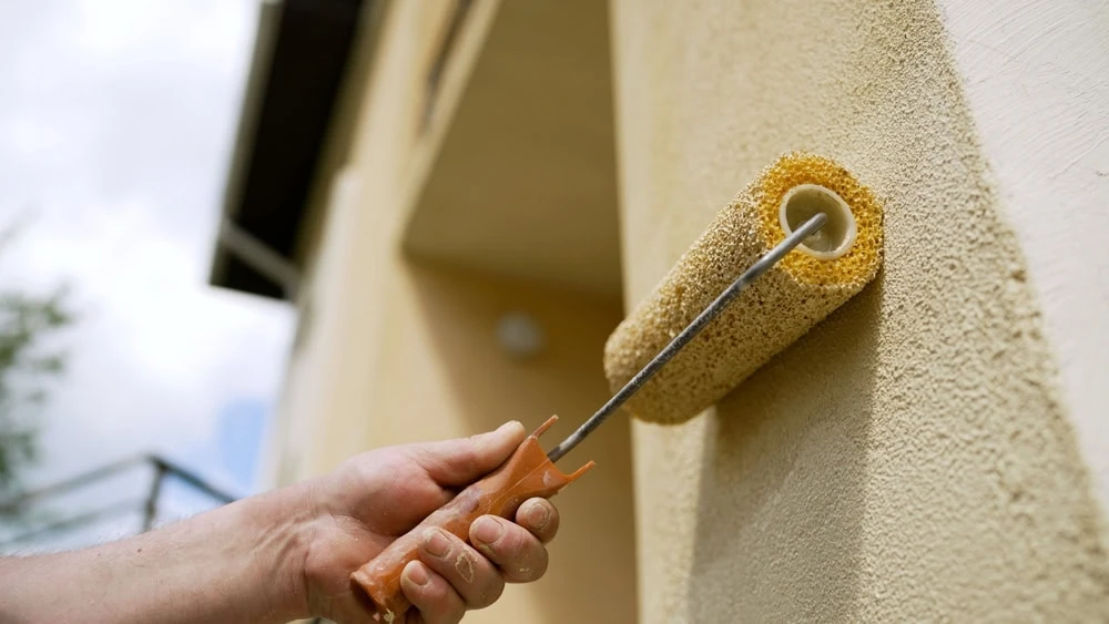 A close-up of a hand using a paint roller to apply light yellow paint to an exterior wall of a building in New Castle County, DE