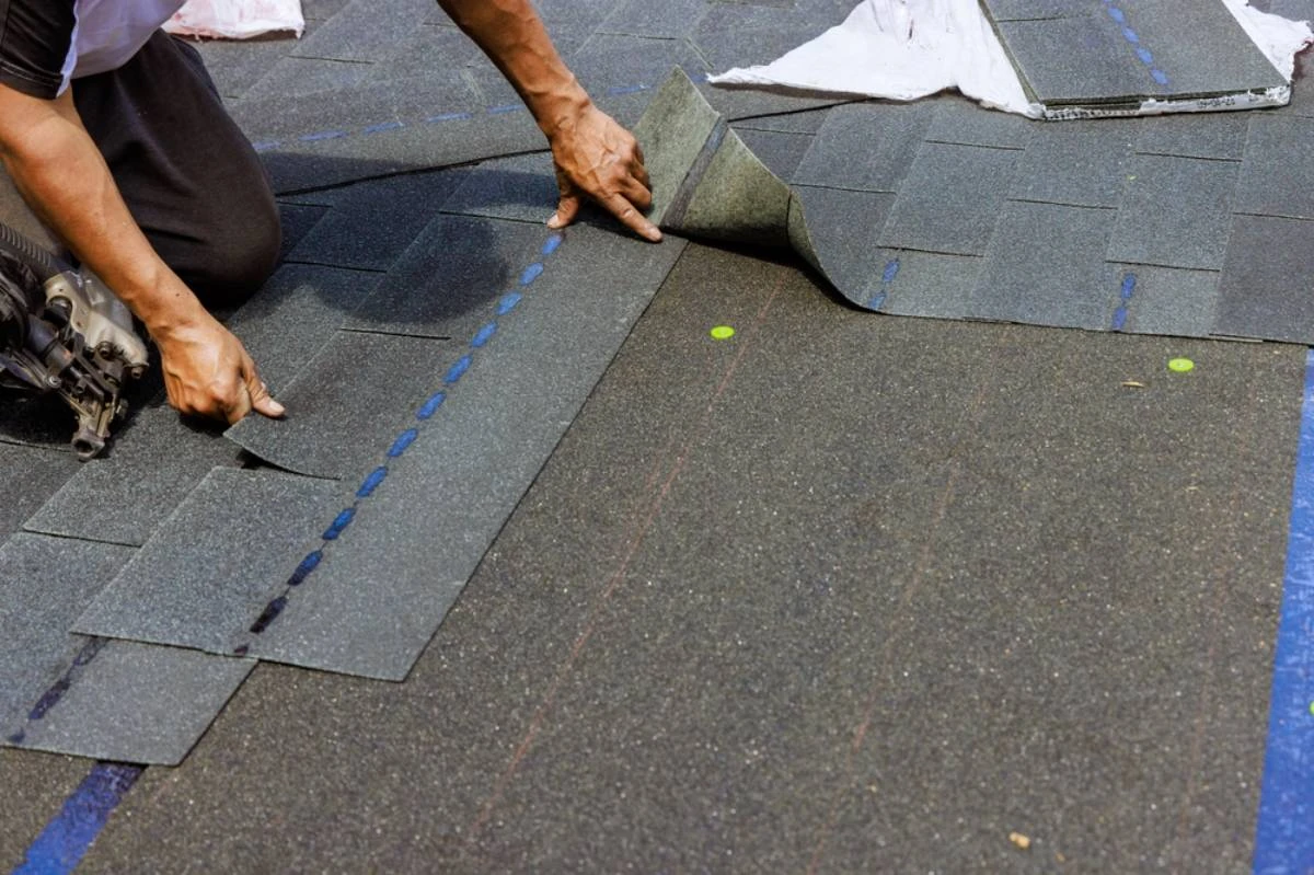 A person installs asphalt roof shingles, positioning and aligning them carefully by hand on a roof. Roofing nails and a roofing nail gun are also visible on the surface in New Castle County, DE