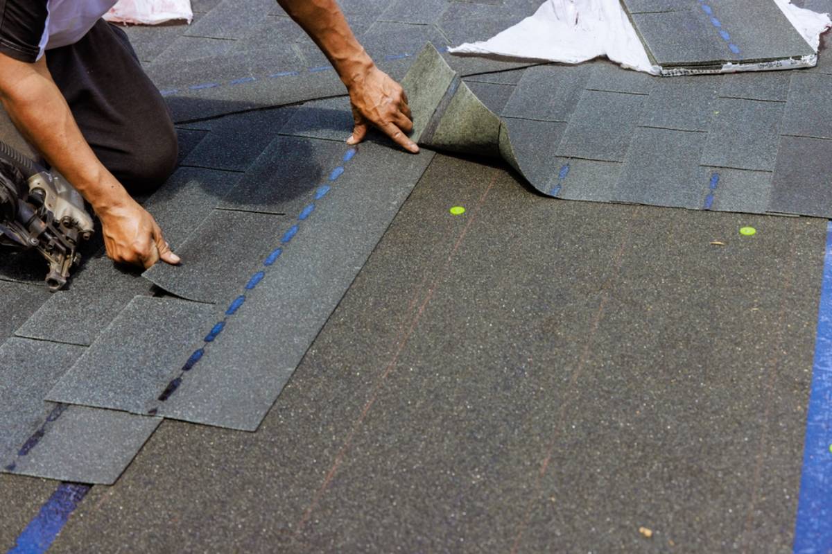 A person installs asphalt roof shingles, positioning and aligning them carefully by hand on a roof. Roofing nails and a roofing nail gun are also visible on the surface.