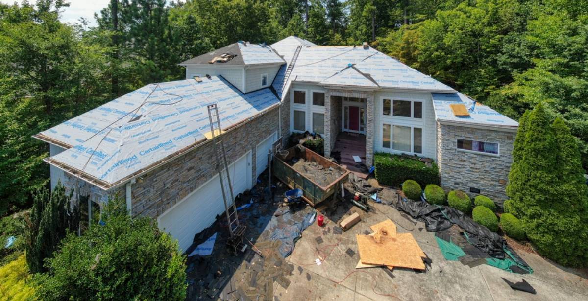 A large house surrounded by trees is having its roof replaced; roofing materials and tools are scattered around, with part of the roof covered in synthetic underlayment and shingles removed.
