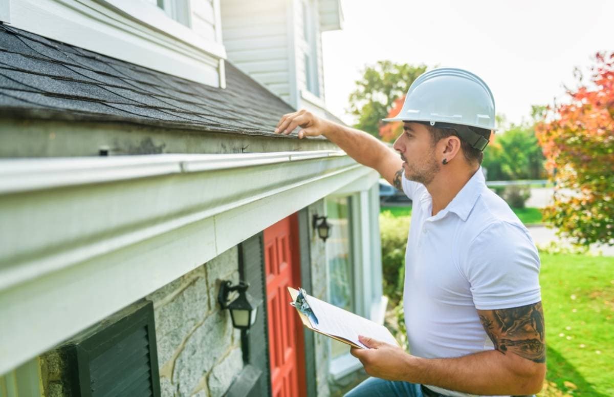 A person wearing a hard hat and holding a clipboard inspects the roof shingles and gutter of a house near the front door on a sunny day.