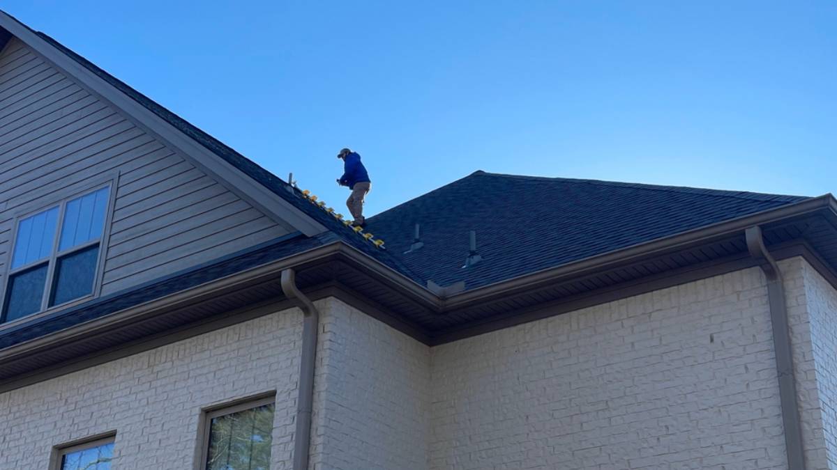 A person stands on the roof of a two-story house, cleaning leaves from the gutter under a clear blue sky.