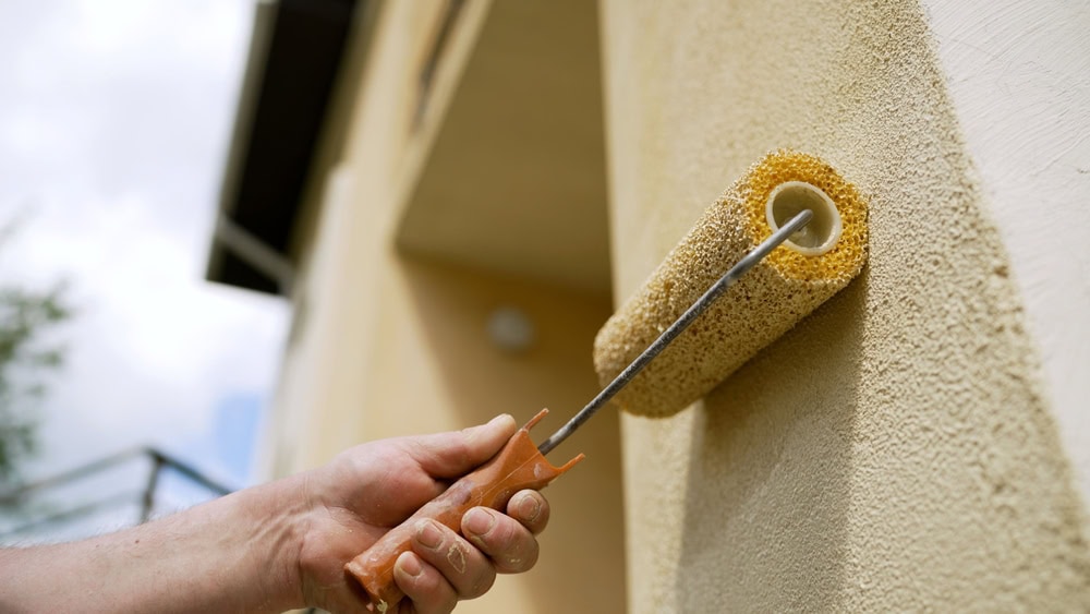 A close-up of a hand using a paint roller to apply light yellow paint to an exterior wall of a building.