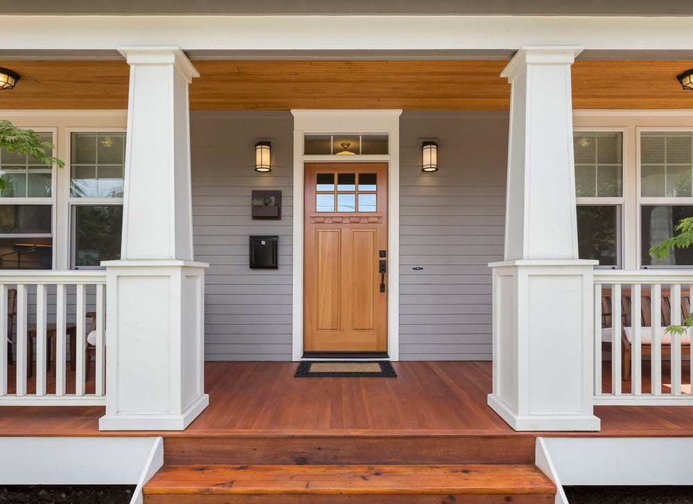 Front porch of a house with wooden steps and flooring, white railings and columns, a natural wood front door with glass panes, two wall lanterns, and light gray siding.